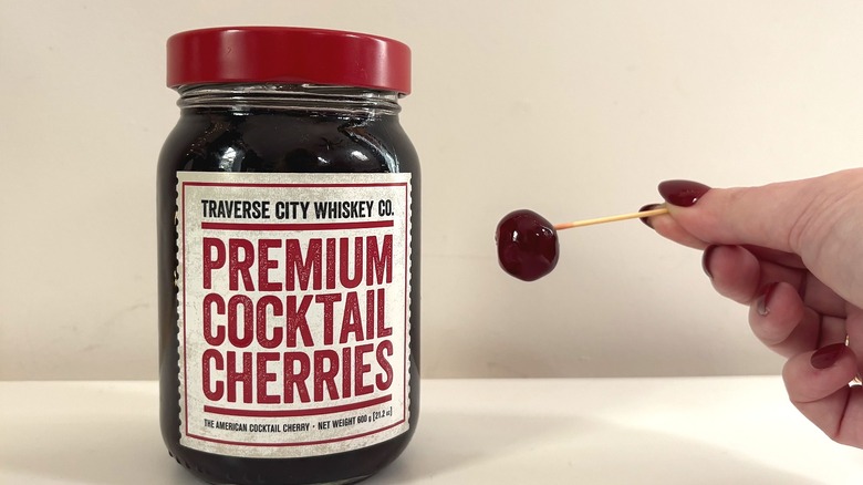 Jar of Traverse City cocktail cherries next to hand holding cherry on toothpick