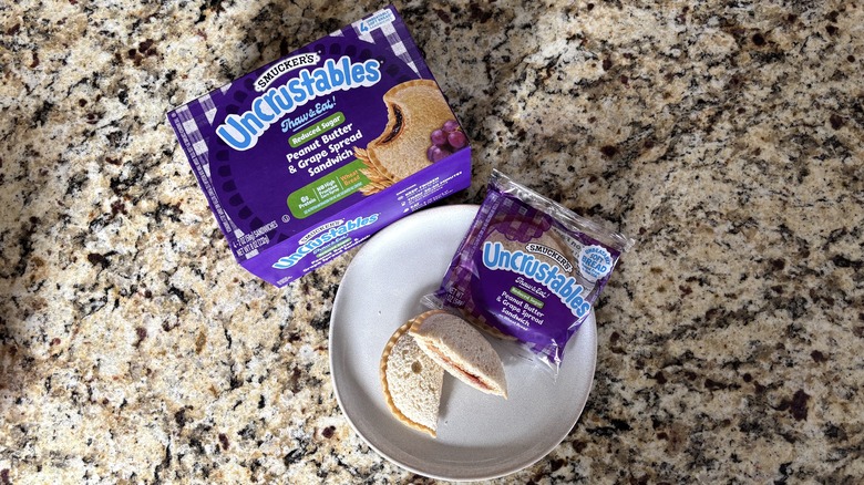 Peanut butter and grape reduced sugar Uncrustables in box, package, and on plate on marble countertop