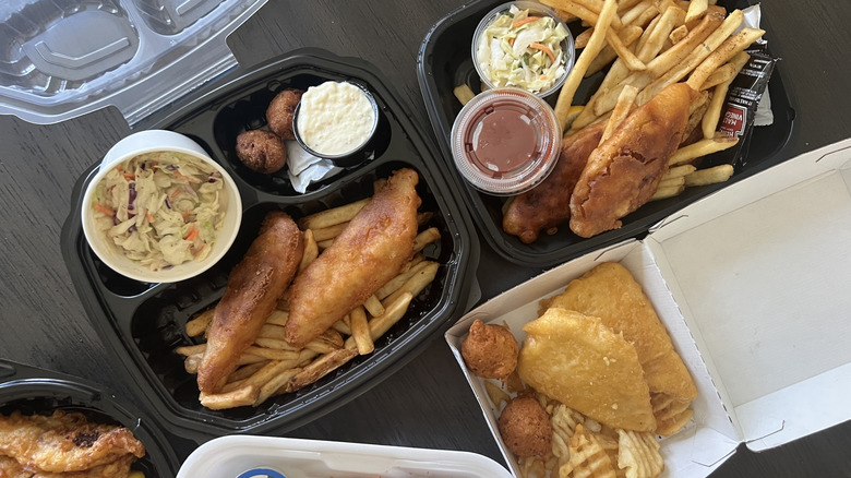 Multiple chain restaurant fish and chips meals in to-go boxes assembled on a dark wooden counter