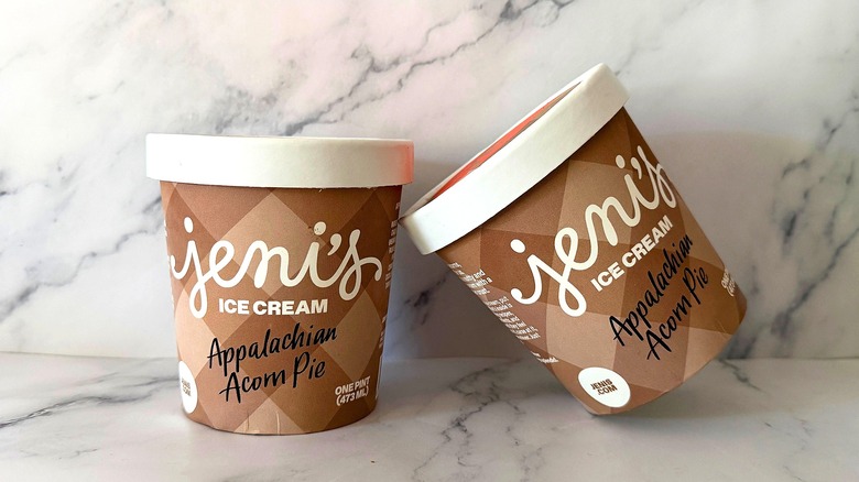 One pint of Jeni's Appalachian Acorn Pie ice cream leaning against another pint of ice cream, all on marble surface
