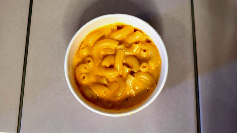 A white takeout container filled with yellow macaroni and cheese on a white tile table.