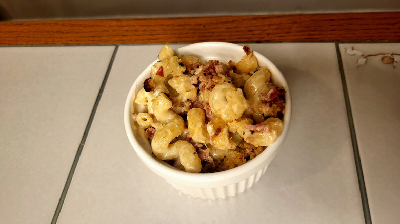 A ramekin full of cavatappi pasta with a white cream sauce, bacon, and breadcrumbs on a white tile table.