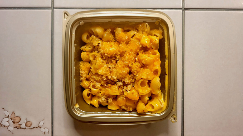 A rounded square metallic takeout container filled with macaroni and cheese and topped with toasted breadcrumbs.