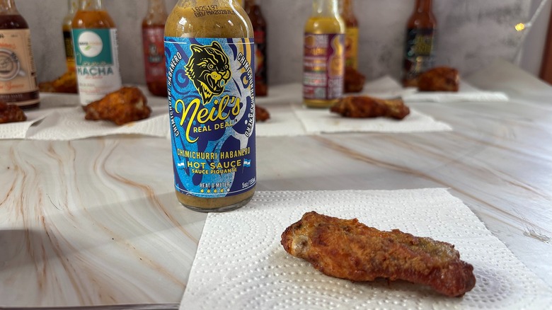Bottle of Neil's Real Deal Chimichurri Habanero hot sauce on table next to chicken wing