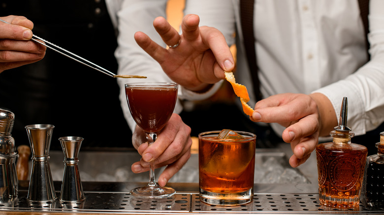 Two bartenders preparing drinks, one garnishing a cocktail with an orange twist and the other adding a dried citrus wheel to the top of a cocktail with culinary tongs