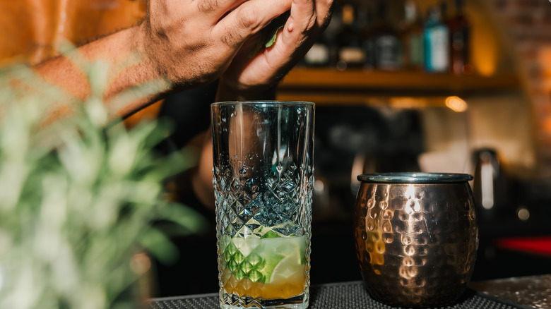 A bartender's hands adding herbs to a glass with limes and other syrups to prepare a cocktail