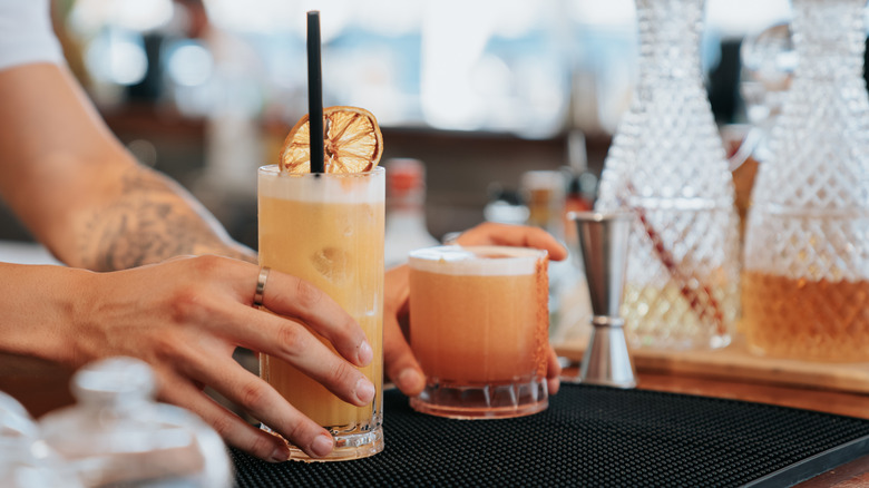 A bartender presenting two cocktails in different sized glasses next to some bartending equipment and decanters