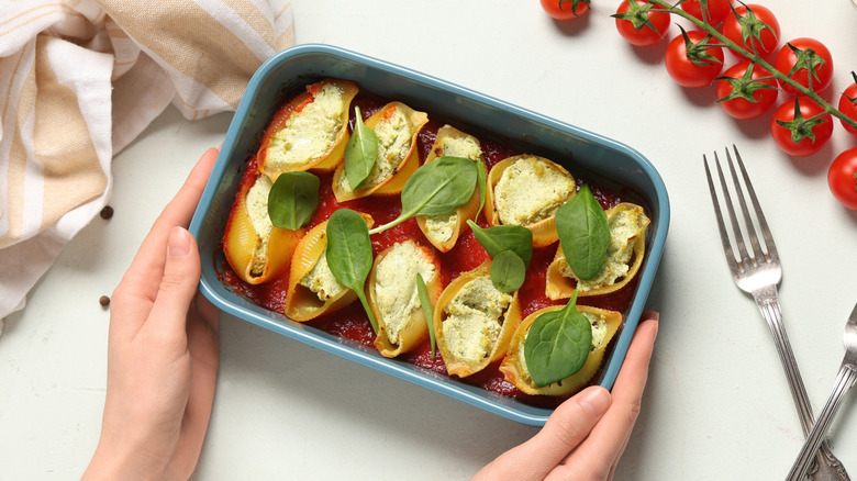 Hands holding baking dish filled with stuffed pasta shells next to forks and ingredients