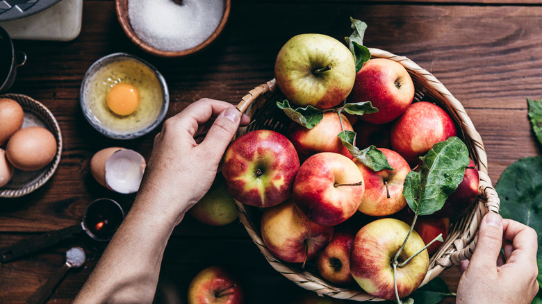 Hands holding basket of yellow and red apples next to cracked eggs and bowl of sugar