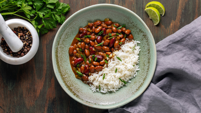Rajma and white rice in a stone bowl on a wood table, surrounded by cilantro, lime slices, and a mortar and pestle filled with peppercorns.