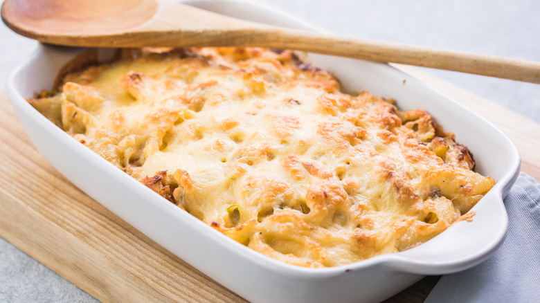 a ceramic dish of baked pasta with a wooden spoon resting on top of it sitting on a wooden cutting board