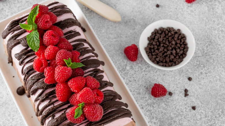 An ice cream cake drizzled with shell topping and topped with fresh raspberries and mint next to a bowl of chocolate chips