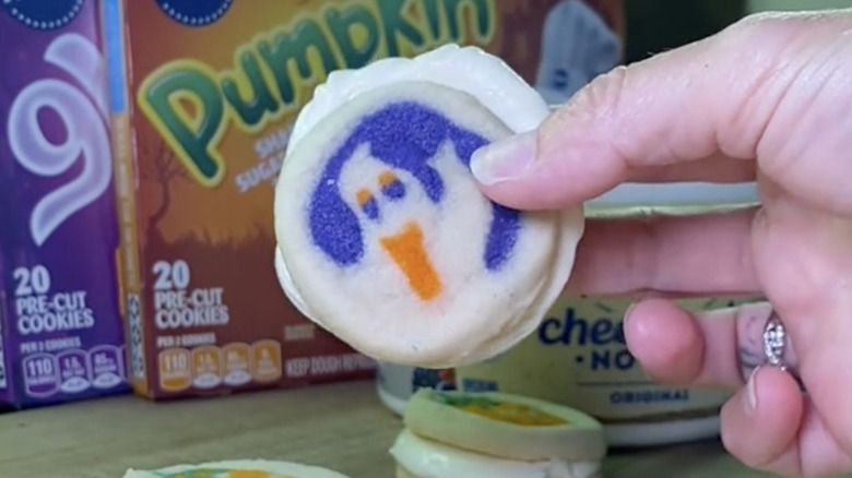 Hand holding a Pillsbury cookie cheesecake sandwich with Pillsbury cookie boxes in the background