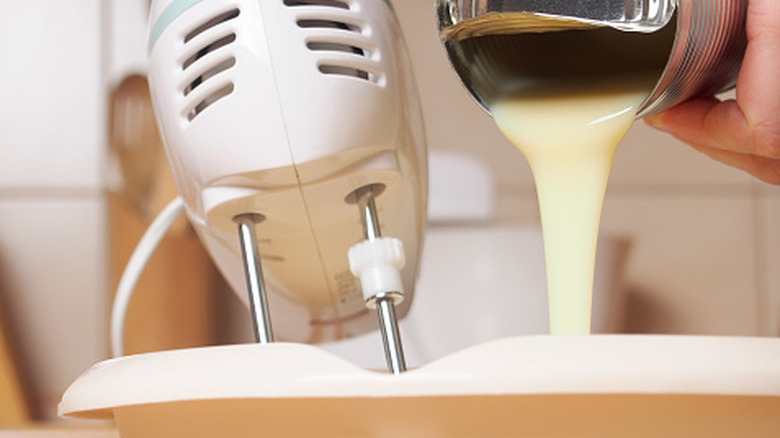 Someone pouring condensed milk into a mixing bowl while holding an electric beater