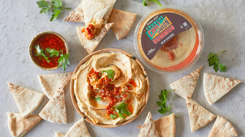 A bowl and a container of Trader Joe's crunchy chili onion hummus surrounded by pita chips and some cilantro