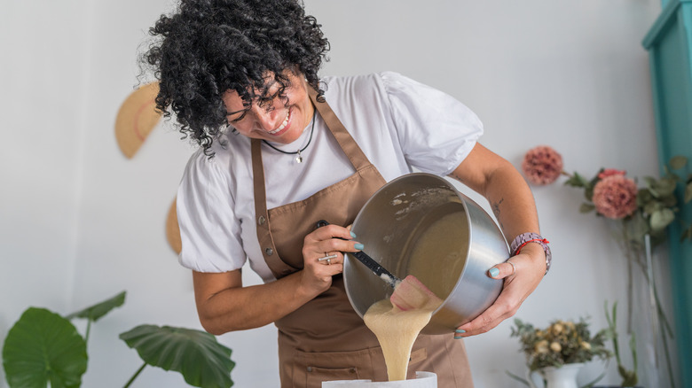 Woman with dark curly hair wearing apron and pouring batter into cake pan