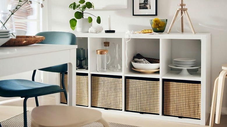 A white KALLAX shelf unit used as sideboard storage in a bright dining area, holding dishes, glassware, and woven baskets, with decorative items and framed art arranged above.