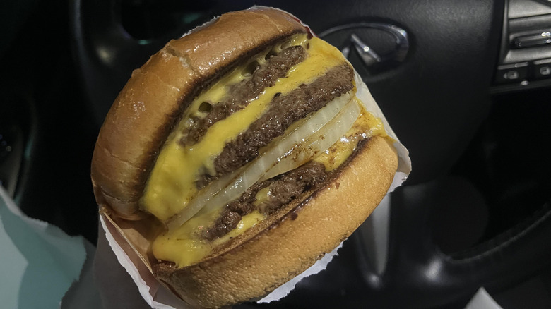 an In-N-Out 4x4 burger with onion being held in front of a car's steering wheel