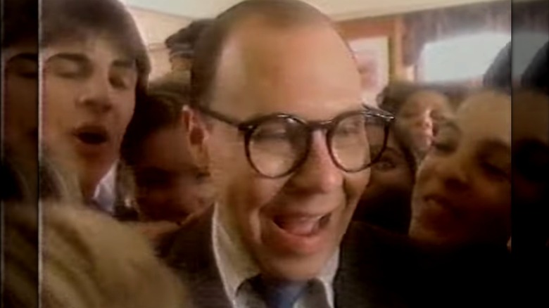 A screen cap of herb, a man with large glasses and a suit adjusting his tie as he enters a Burger King