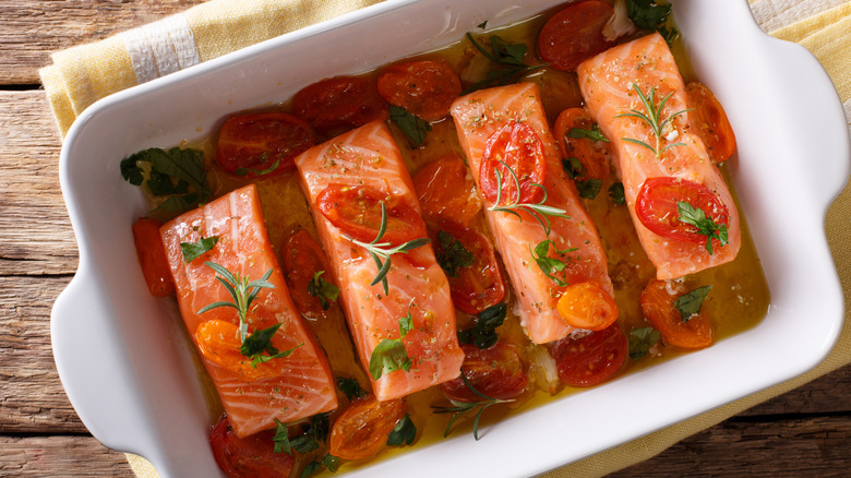 White baking dish holding four salmon filets with roasted cherry tomatoes, rosemary, and green hervs