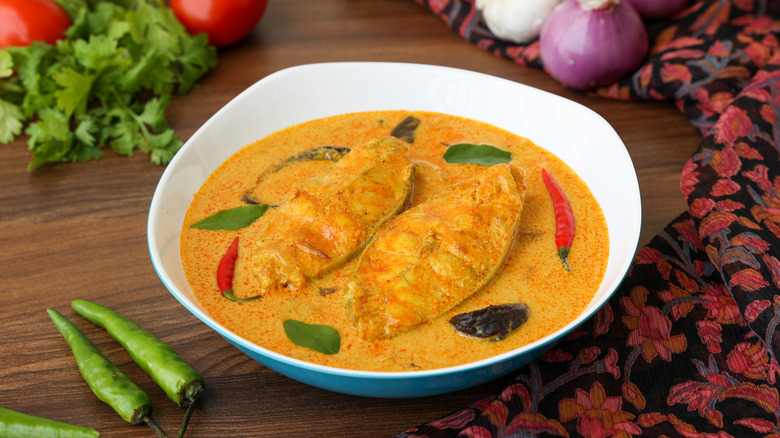 Goan fish curry in bowl surrounded by onions, cilantro, green chilies, and tomatoes