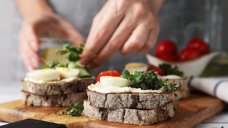 A person's hands assembling a sandwich with eggs, cheese, condiments, bread, and tomatoes