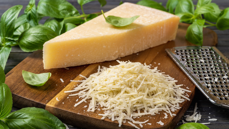 Block of whole Parmesan on a wooden board with some gratings and herbs on the side