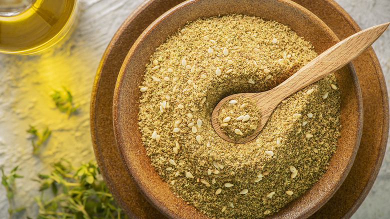 Top view of a bowl of za'atar with a wooden spoon in it next to a bottle of oil