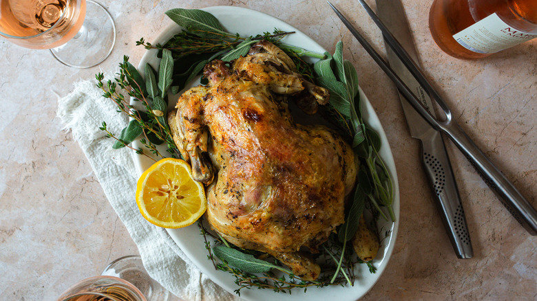 whole chicken on platter with herbs, lemon, carving tools and wine