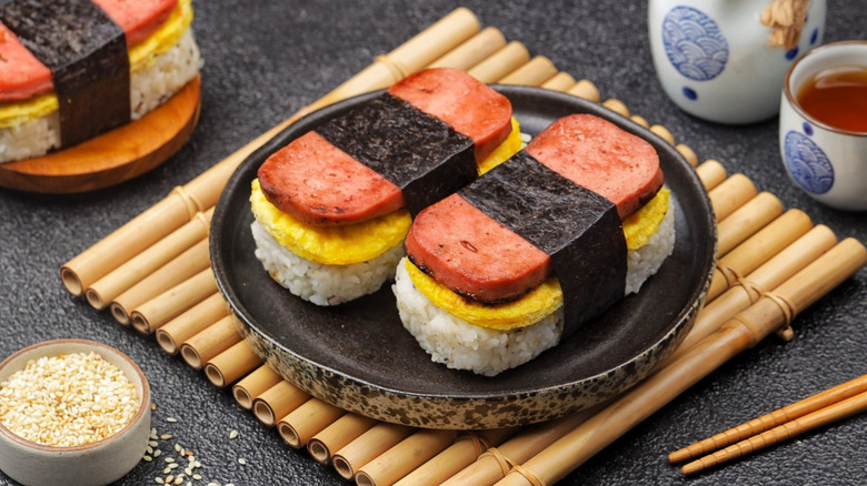 Spam musubi on black plate with chopsticks, soy sauce, and sesame seeds