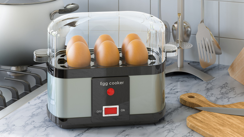A gray and black egg cooker with a red on/off button, standing on the counter, holding six eggs