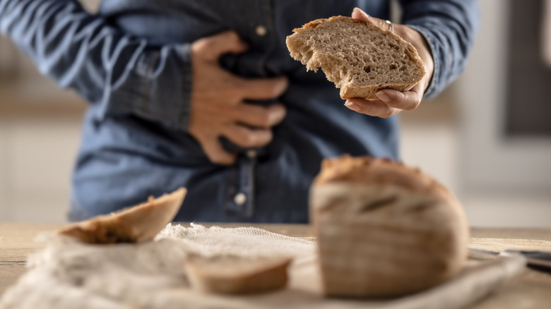A person clutches their stomach, and a piece of bitten bread, in front of a cutting board with sliced bread
