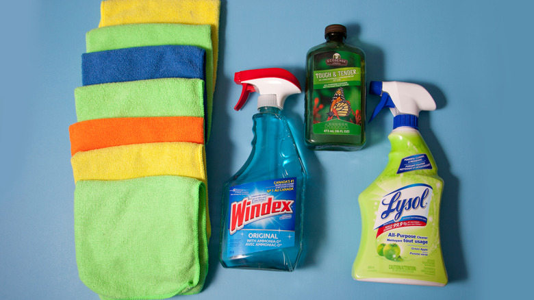 An array of household cleaning products, including microfiber cloths, Windex, Lysol disinfectant spray, and Tough &  Tender