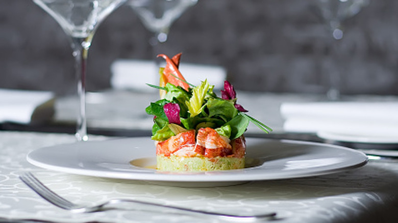 A seafood dish at an upscale restaurant delicately plated on a white dining plate, on linen tablecloths, beside a fork and several wine glasses