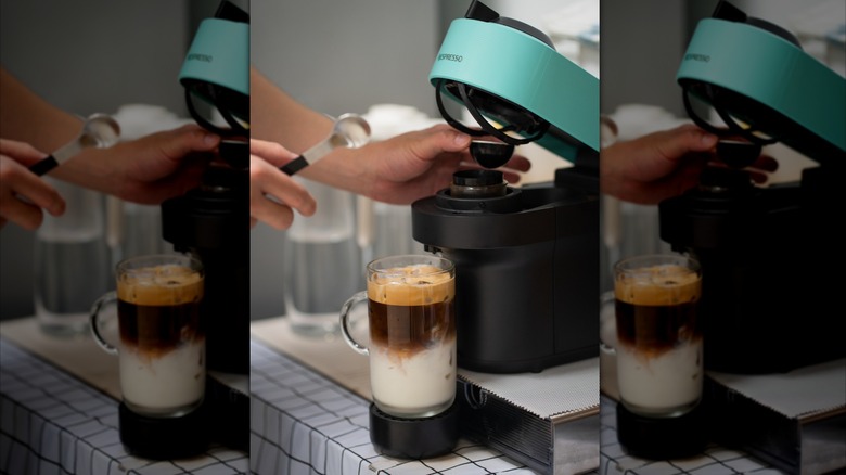 Someone changing the pod on a blue Nespresso machine, with a glass mug of coffee and milk.