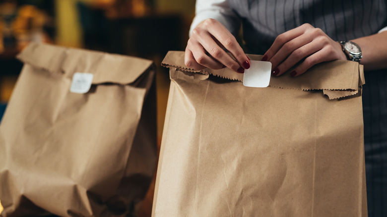Manicured hands packing up a to-go order of food into brown paper bags that are sealed shut with a white sticker.