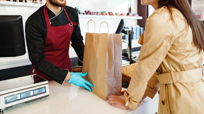 A customer grabbing a food order in a brown paper bag from an employee on the other side of the counter with a red apron and blue plastic gloves on.