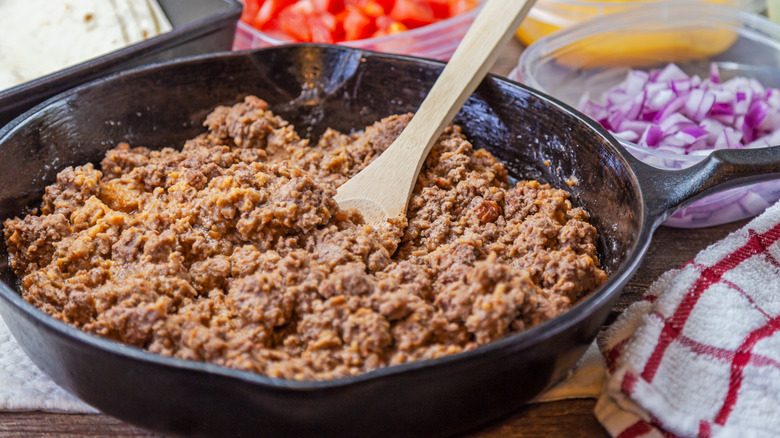 A skillet of browned ground beef with a wooden spoon in it and tomatoes and onions in the background