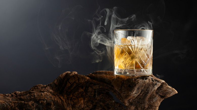 A glass of whiskey on the rocks with smoke around it sitting on a strange piece of wood