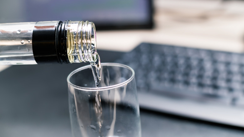 A bottle of vodka pouring into a clear glass in front of a computer keyboard on a desk in the background