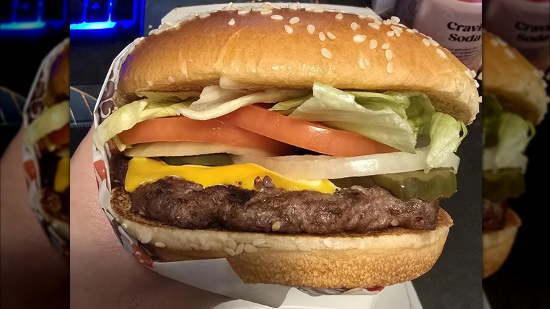 A Whopper wrapped in paper, on a red fast food tray, with an order of fries and a packet of ketchup, and a drink is in the background