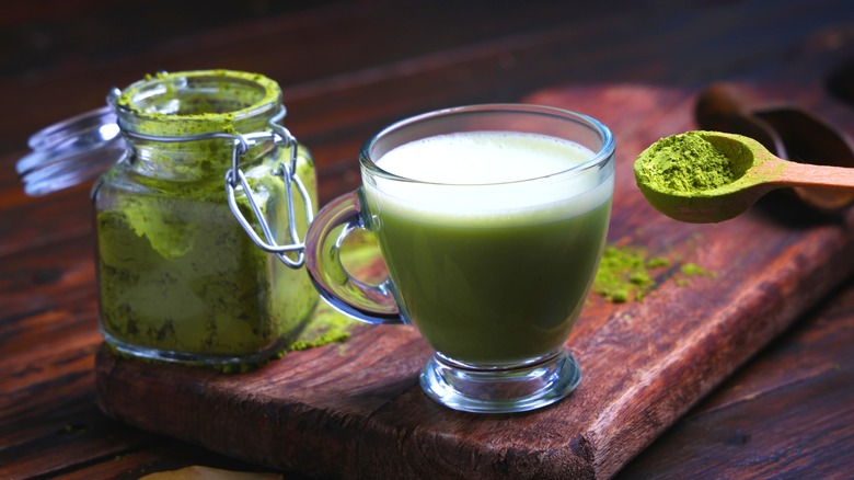 Open glass jar and wooden spoon full of matcha powder next to cup of brewed matcha on wooden board