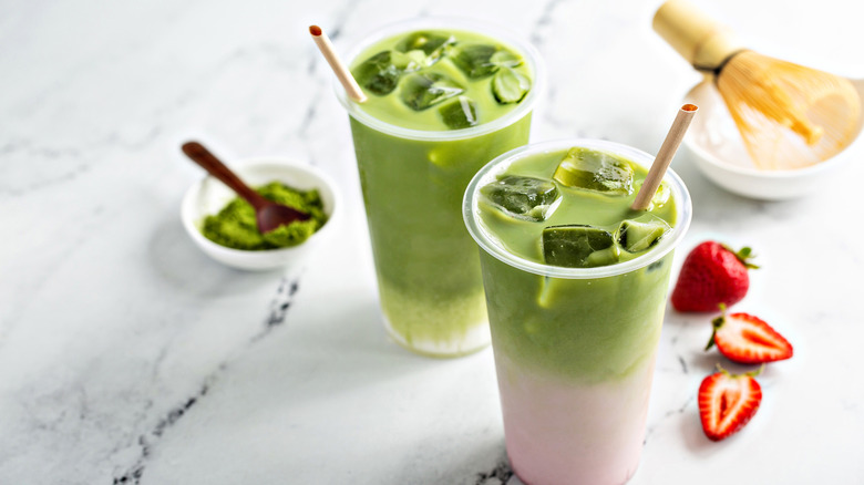 Iced strawberry matcha latte and iced plain matcha latte in tall glasses with straws next to strawberries and matcha powder