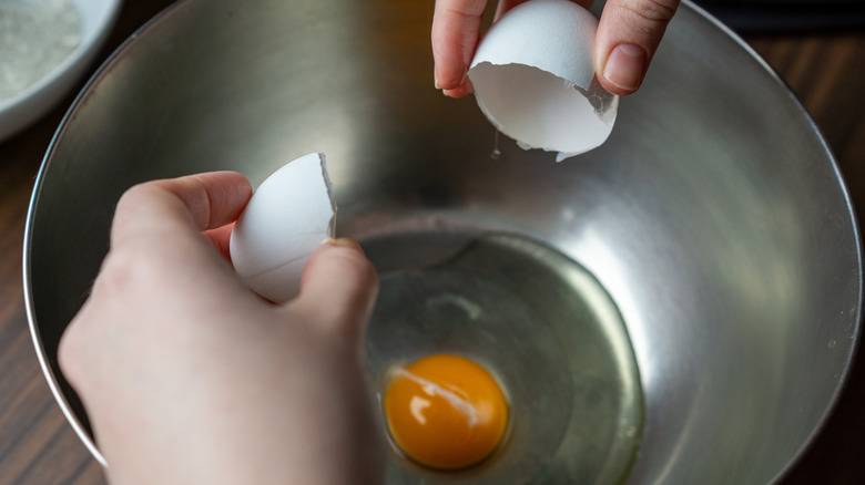 A person cracks and egg into a metal bowl