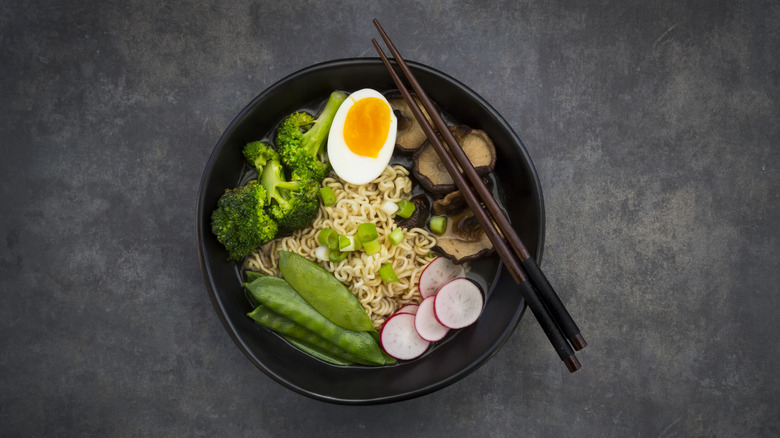 Ramen with sugar snap peas, egg, broccoli, mushrooms and radish in a black bowl with black chopsticks - birds eye view on a black table