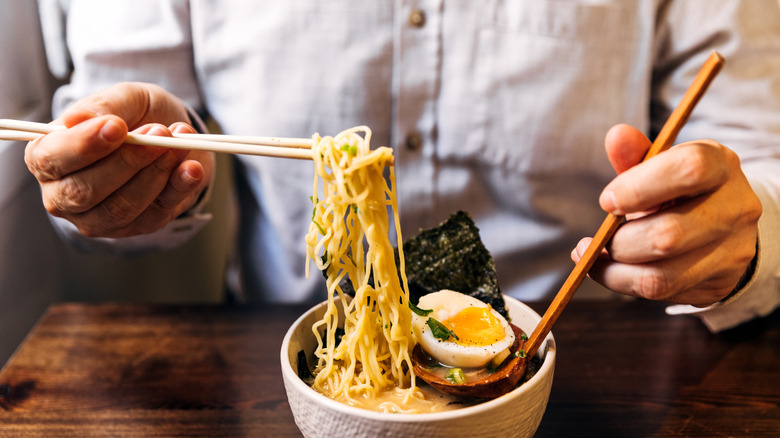Close up of a person eating ramen with chopsticks and a spoon, white bowl place on a wooden table