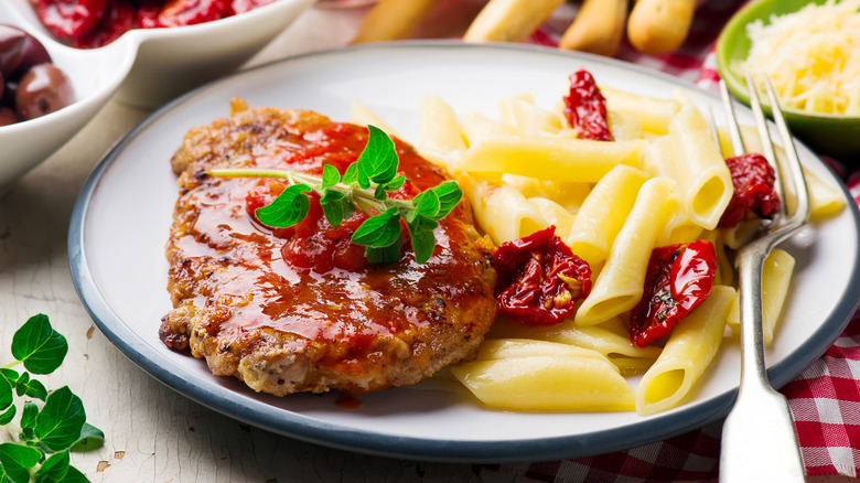 A pork chop alla pizzaiola with a side of pasta and sun-dried tomatoes on a white plate with a fork, with a bowl of grated cheese in the background