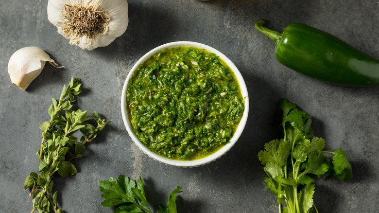 Tangy chimichurri sauce in a white bowl with ingredients surrounding it like garlic, jalapeño and herbs.