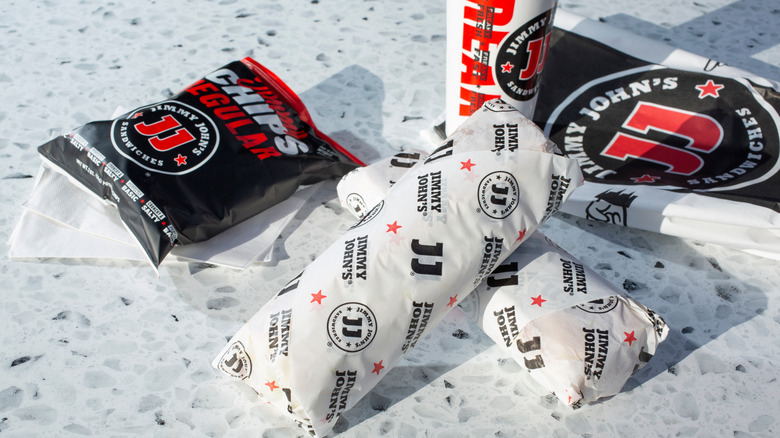 Two wrapped Jimmy John's subs with chips, a beverage cup, and a bag on a marble table
