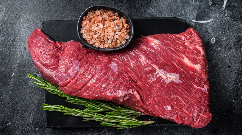 Large raw steak on a cutting board beside rosemary and salt